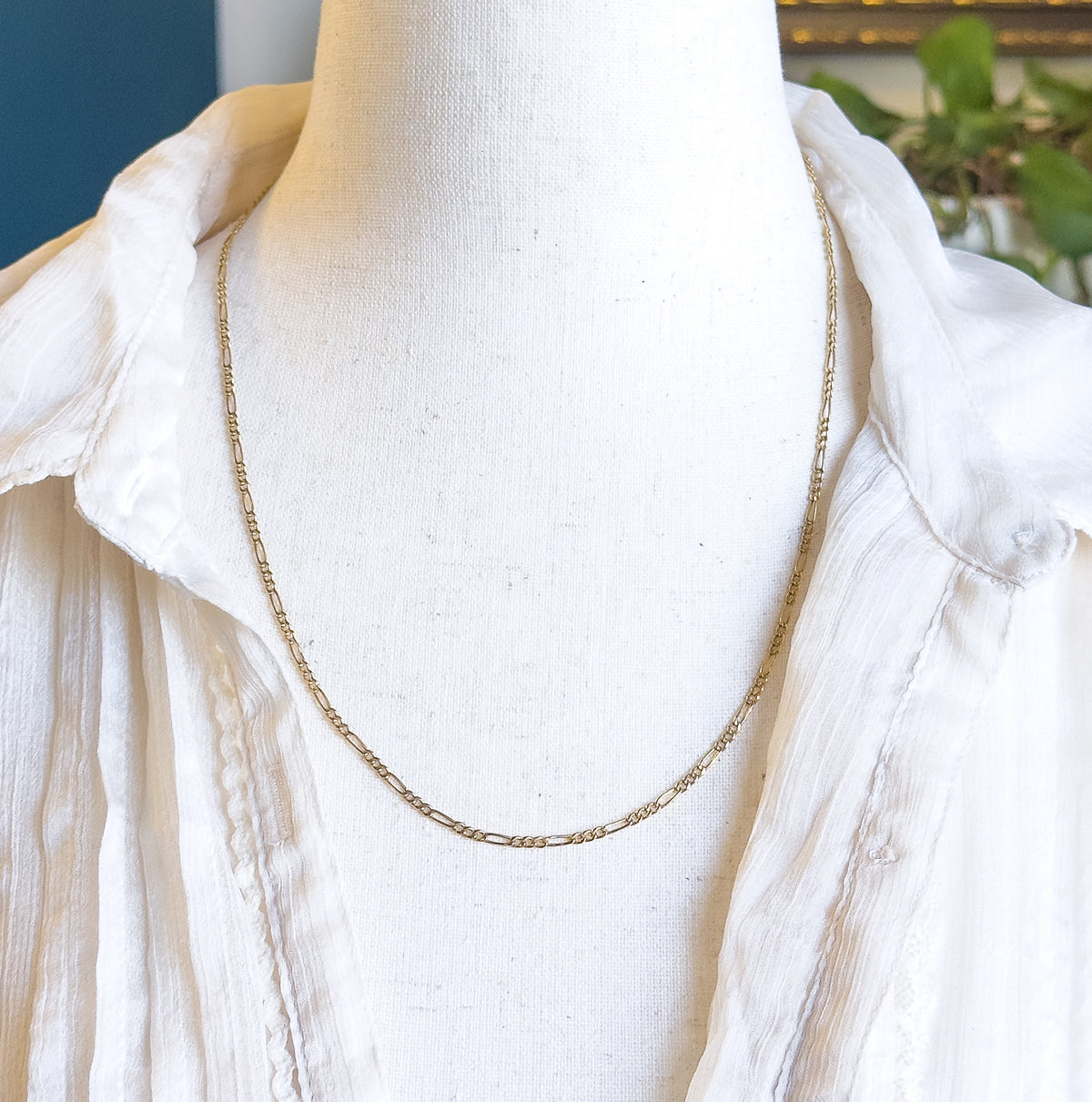 A 14K yellow gold Figaro link chain necklace, shown at the neck of a cream fabric bust form dressed in an open white blouse, its alternating short and long links catching the light.