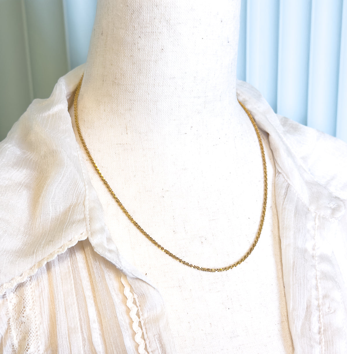 The chain shown at the neck on a cream linen dress form in an open-collared blouse, the fine textured links tracing a slim curve to the collarbone.
