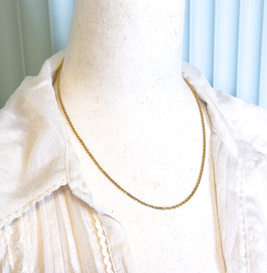 The chain shown at the neck on a cream linen dress form in an open-collared blouse, the fine textured links tracing a slim curve to the collarbone.