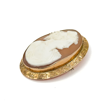 Tilted three-quarter view against plain white, the oval standing on its side so the engraved floral gold border catches the light along the near edge and the pin stem tip shows behind.