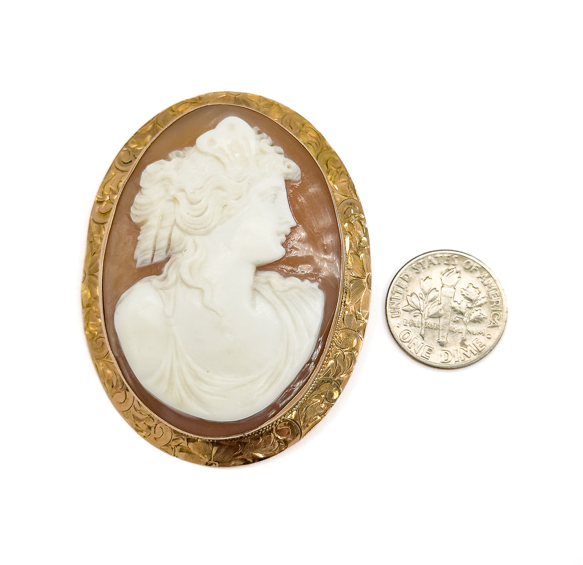 Face-up against white beside a dime, the coin reaching only about a third of the oval's height to show how large the shell cameo and engraved gold mounting are.