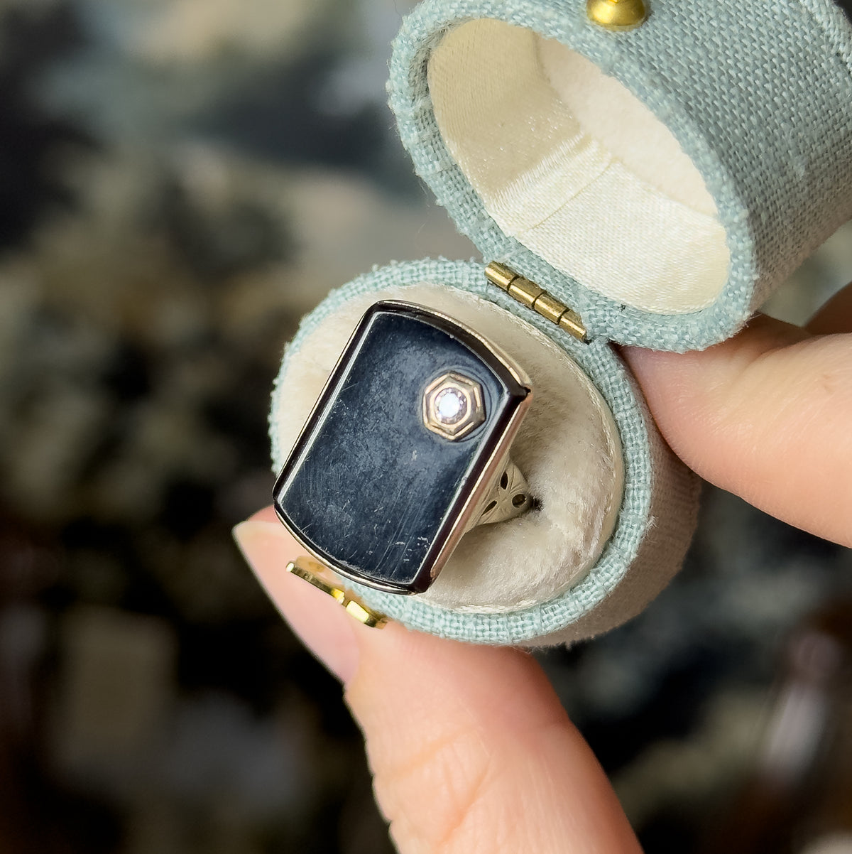Art Deco onyx and diamond ring in 10K white gold, shown tucked inside a small pale green ring box held between finger and thumb, its long black bezel-set face centred by a tiny bright stone.