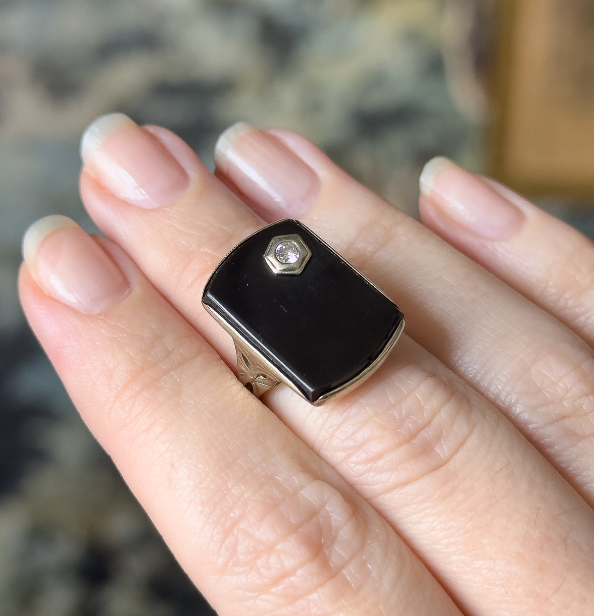 Shown on a finger, the elongated black onyx face spans the width of the finger, its hexagonal bezel and small bright diamond set high, with etched leaf shoulders at each side.