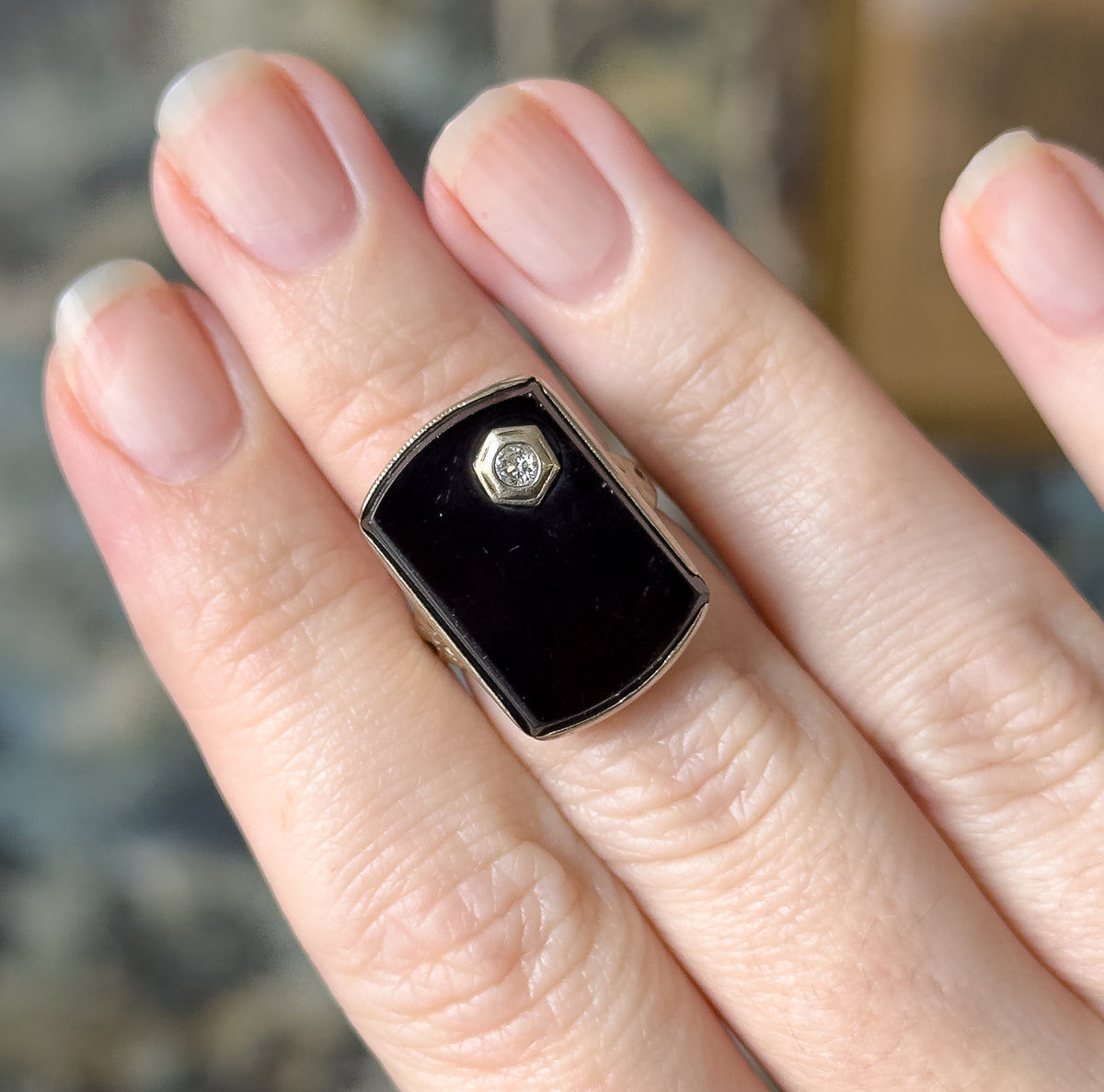 Close overhead view on the hand, the onyx face filling the finger from knuckle to knuckle with the small hexagonal diamond bezel just above centre.