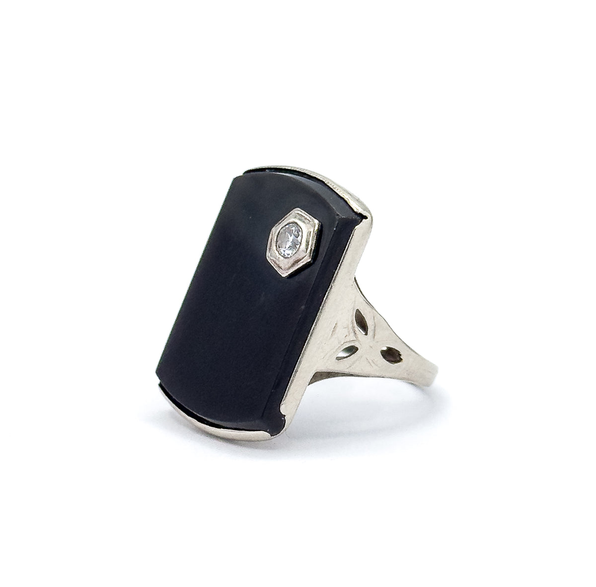 Angled side view against white, showing the onyx face in profile with its white gold bezel edge, the hexagonal diamond setting, and the pierced shoulder with etched leaf cutouts meeting the shank.