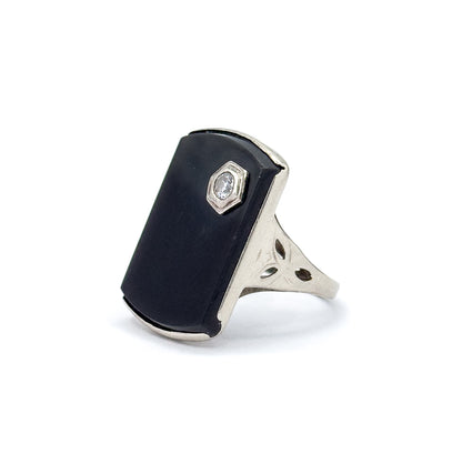 Angled side view against white, showing the onyx face in profile with its white gold bezel edge, the hexagonal diamond setting, and the pierced shoulder with etched leaf cutouts meeting the shank.