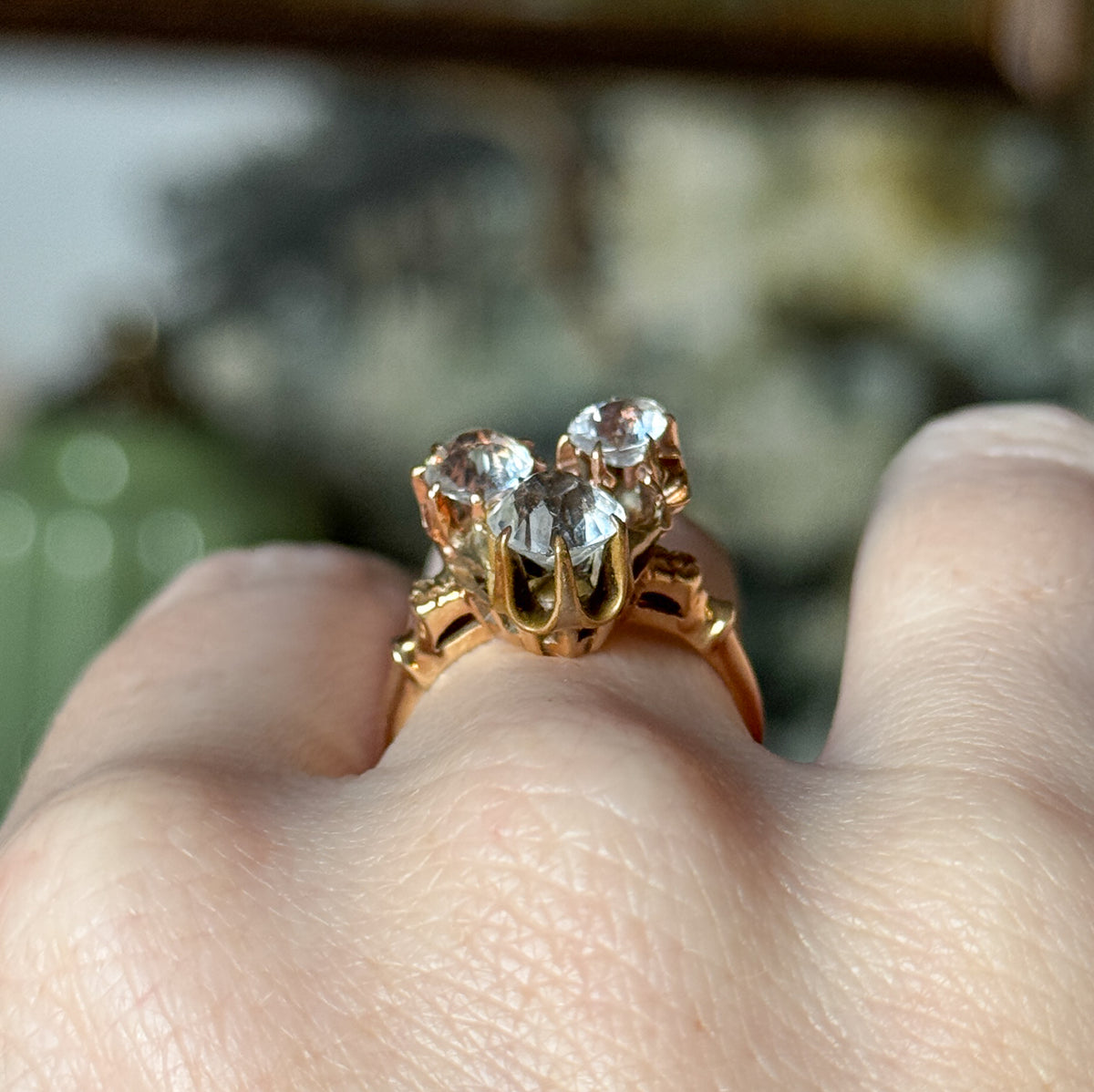 A Retro paste stone ring in 14K yellow gold, shown on a finger, its three colourless stones held in prong baskets above a polished shank with orange blossom detailing along the sides.