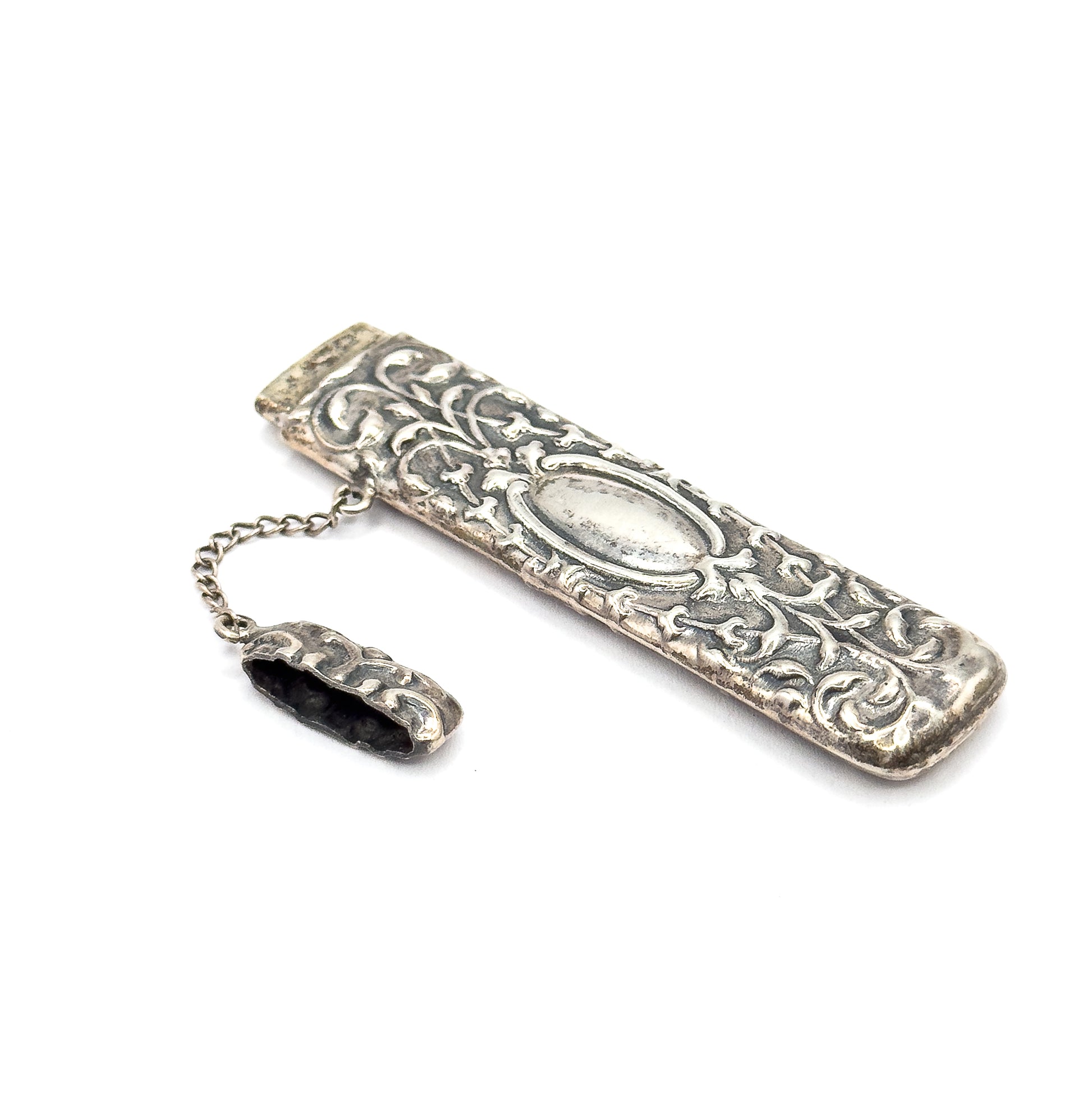 The case lies diagonally on white with the cap detached and resting on its chain, showing the hollow interior opening and the oval panel amid scrolling repoussé.