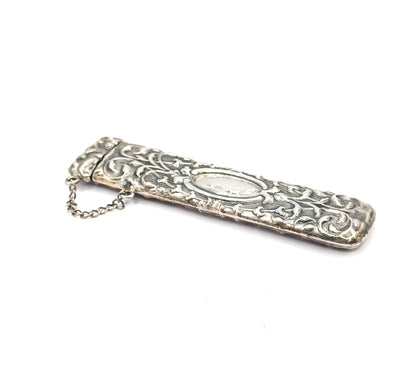 The case seen from above at a steep diagonal on white, cap end nearest the viewer with the chain curving loose beside it, the plain oval panel and darkened patina in the scrollwork clear.
