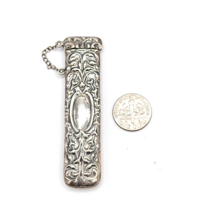 The case stands upright beside a dime for scale, roughly three times the coin's width, its cap and looping chain at top above the floral repoussé and central oval panel.