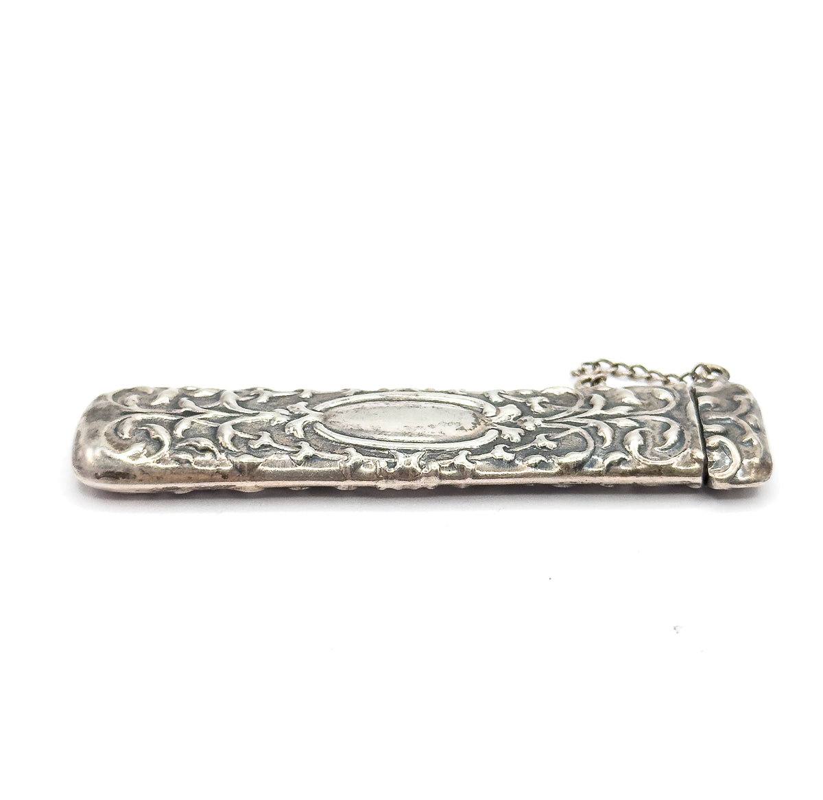 The case lies almost flat and lengthwise on white, viewed low from the side so the cap and its chain sit at the right and the oval panel faces the viewer.