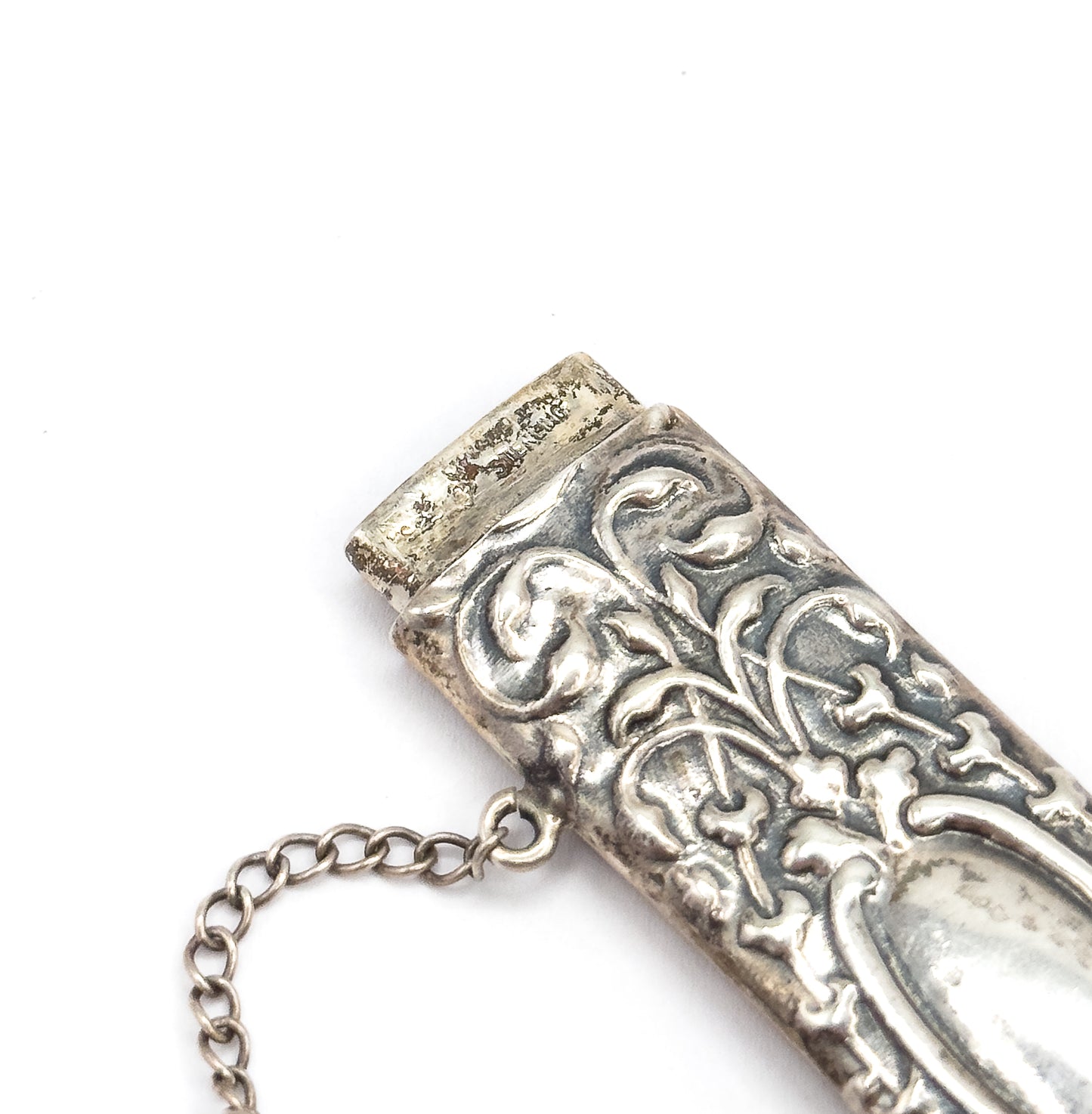 An Edwardian floral repoussé needle case in sterling silver, shown close up at an angle on white, with its removable cap lifted slightly and a fine link chain trailing from the side.