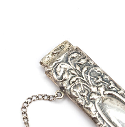 An Edwardian floral repoussé needle case in sterling silver, shown close up at an angle on white, with its removable cap lifted slightly and a fine link chain trailing from the side.
