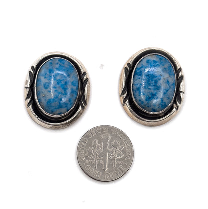 Both earrings face up beside a dime, which shows their scale, with the blue stone cabochons and patinated silver borders clearly visible.