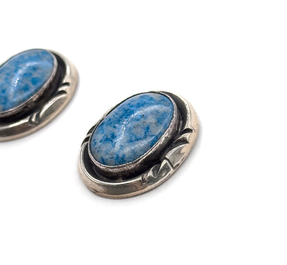 Close crop on one earring tilted towards the camera, filling the frame so the cabochon's blue and pale grey colouring and the leaf-shaped silver detail beside the bezel are clearly seen.