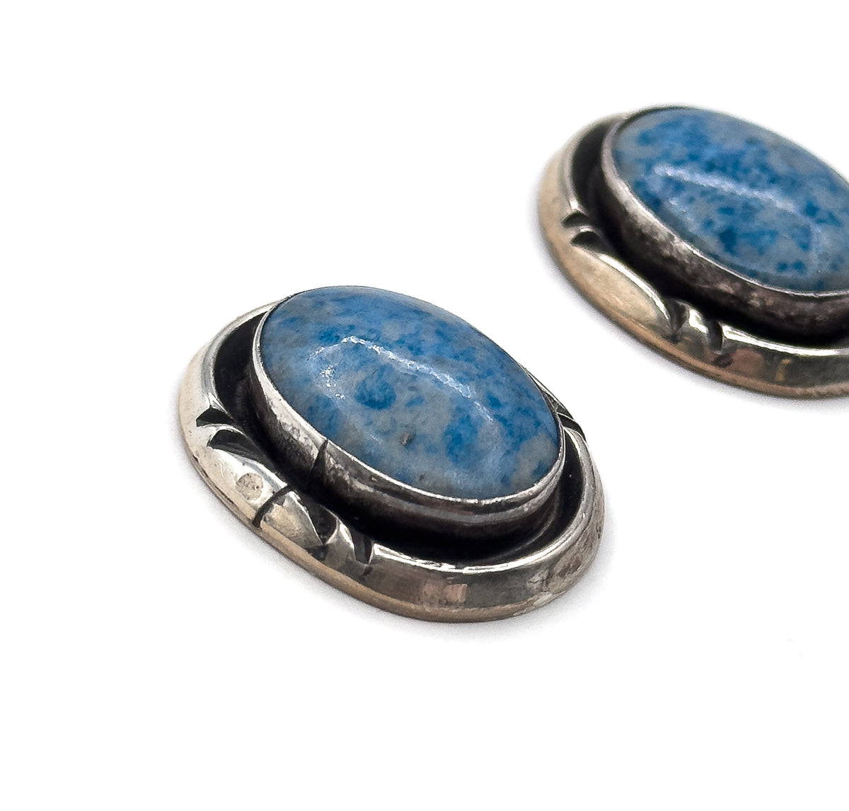 Angled view with the nearer earring in sharp focus and the second softly blurred behind, showing the height of the bezel wall and the domed curve of the blue stone cabochon.