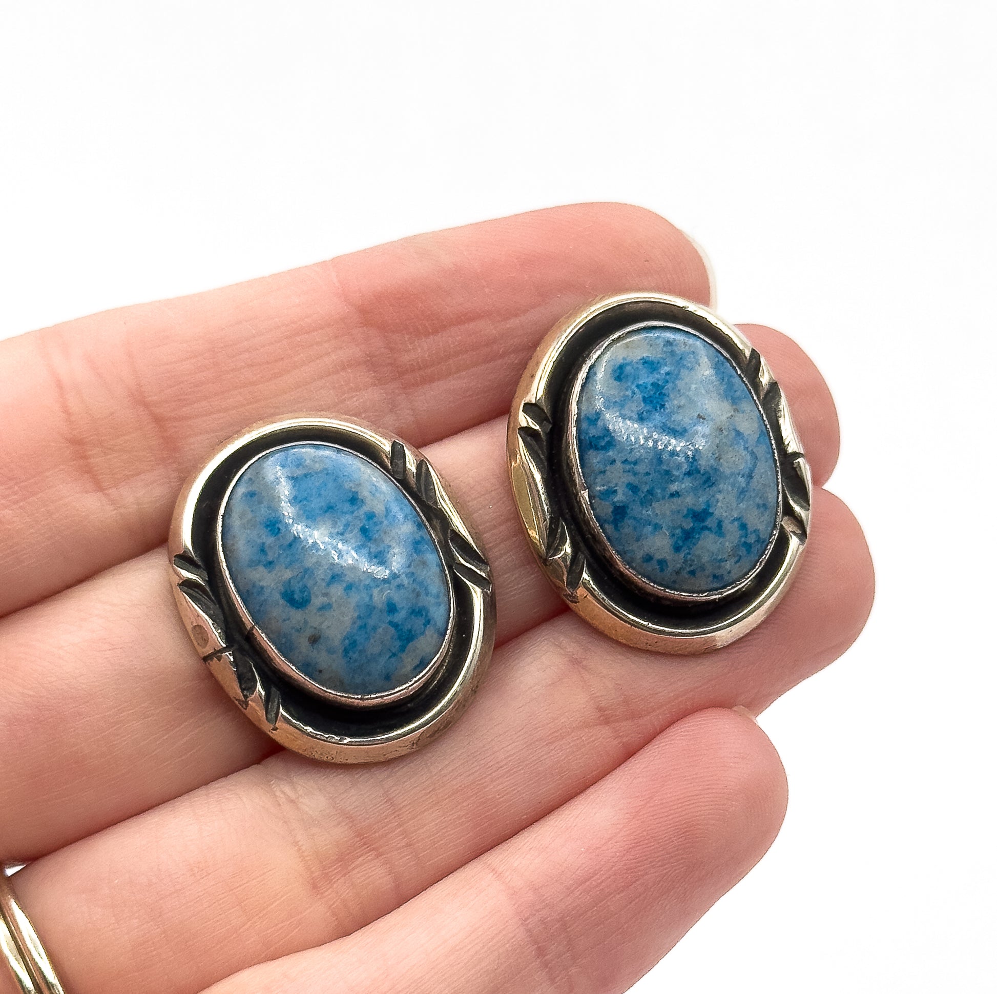 A pair of sterling silver earrings with large oval blue stone cabochons in bezel settings, shown resting on open fingers, which gives a sense of their size, against a white background.