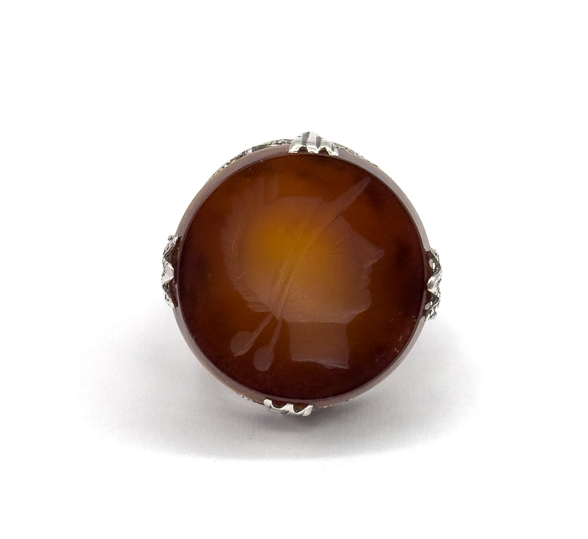 Straight-on top view filling the frame with the round rootbeer quartz, the right-facing soldier intaglio clearly readable between four leaf-shaped sterling silver prongs.
