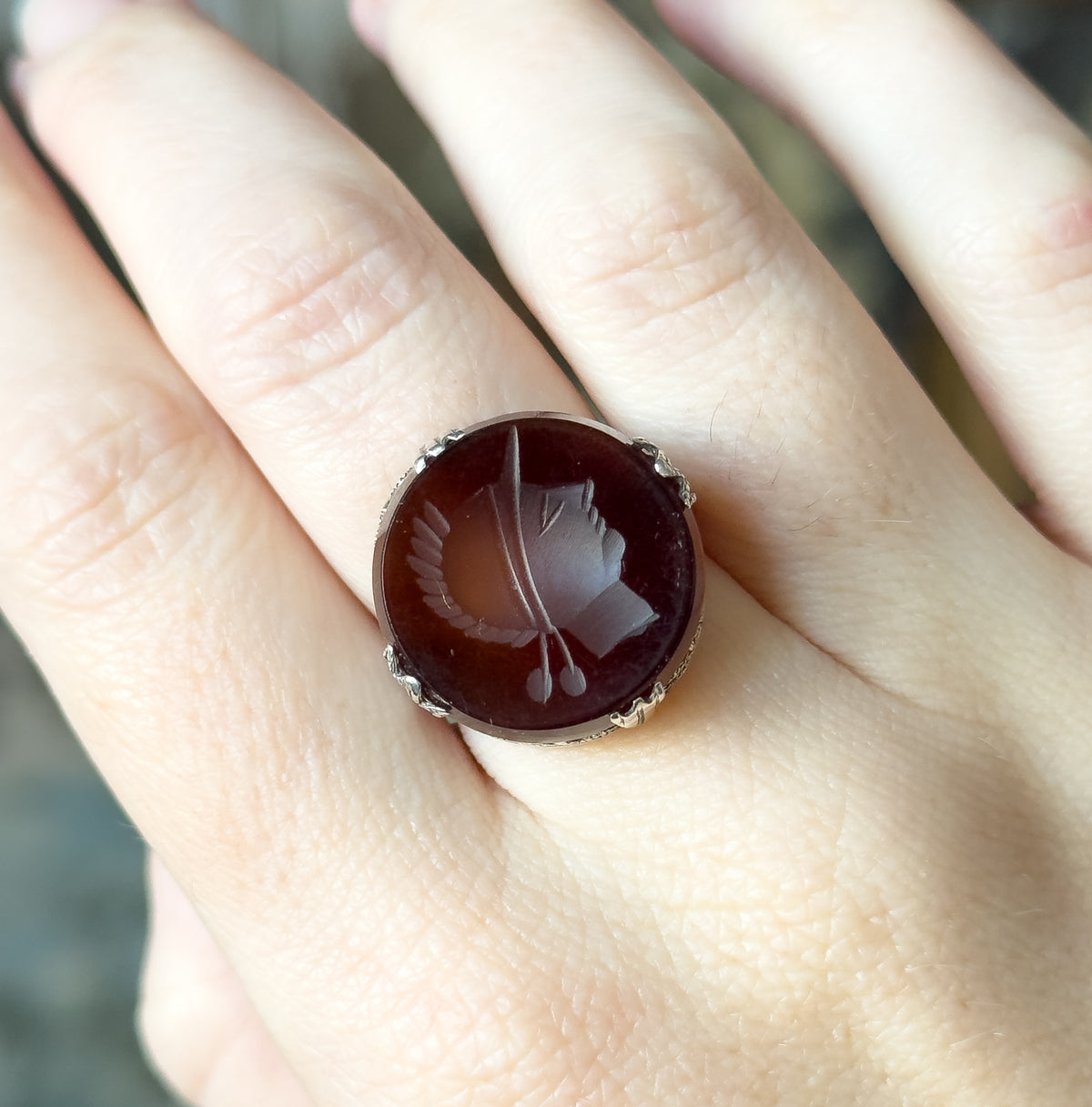 Close overhead view on the hand, the round rootbeer stone spanning almost the full width of the finger, with four decorative prongs gripping its edge.