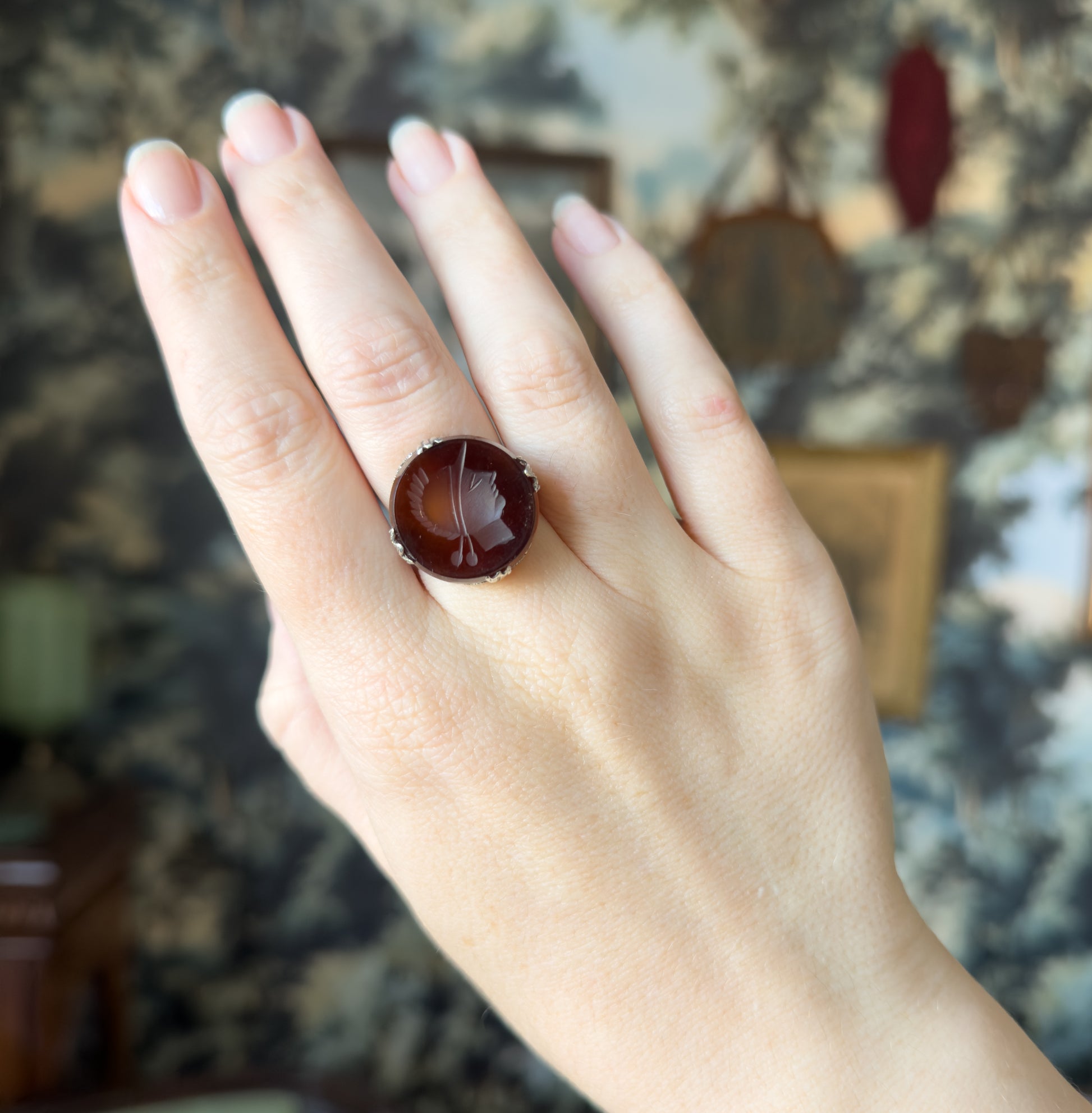 The whole hand raised against a patterned backdrop, the ring shown on the middle finger so its round stone reads clearly against the fingers for scale.