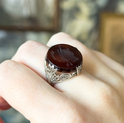 Vintage intaglio carved quartz ring in sterling silver, shown on a finger, the round deep rootbeer stone carved with a right-facing soldier profile above a filigree gallery and prongs.