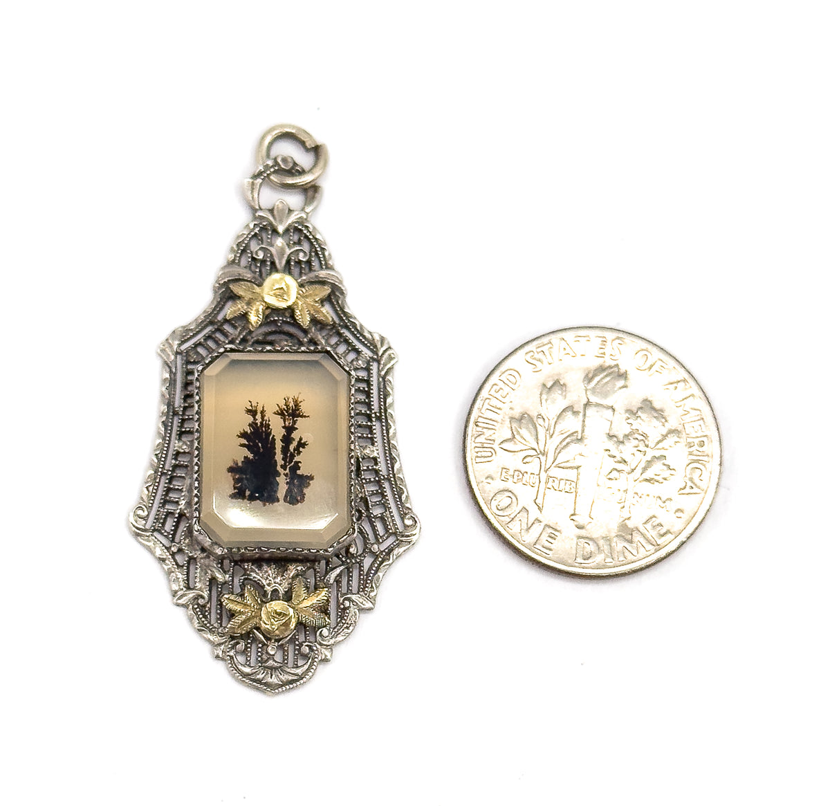 Laid flat beside a dime for scale, the pendant stands a little taller than the coin, its jump ring bail at top and the dark fernlike markings clear in the pale bezel-set stone.