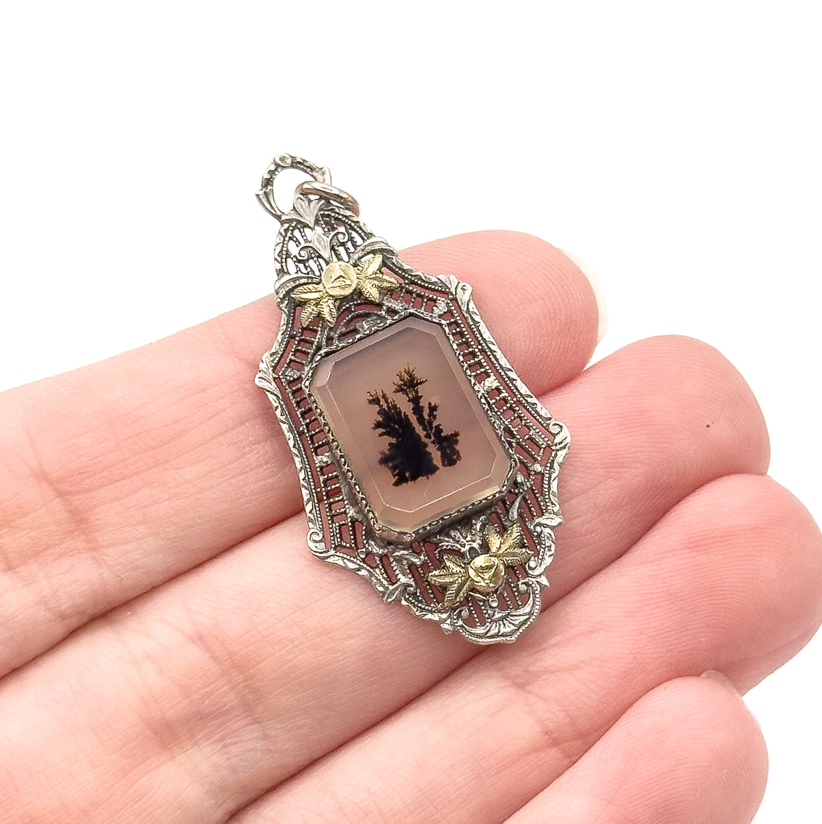 Art Deco dendritic agate filigree pendant in sterling silver, shown resting on open fingers for scale, its bezel-set stone framed by openwork and gold floral accents above and below.