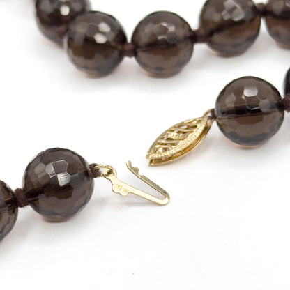 The clasp shown open at last, its pierced gold oval on one bead end and the hooked tongue with a tiny stamp on the other, lying apart on white between the faceted brown beads.