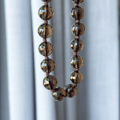 A vintage smoky quartz opera necklace of round faceted deep brown beads, hand knotted between each bead, shown hanging in two strands against pale draped fabric.