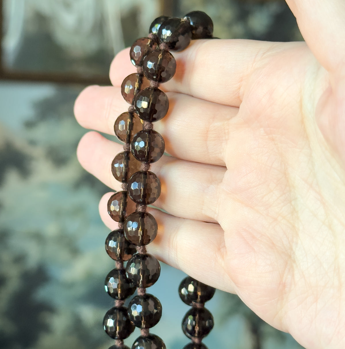 The faceted smoky quartz beads draped over a hand, fingers behind the strand giving a sense of scale, with the knots between beads clearly visible.