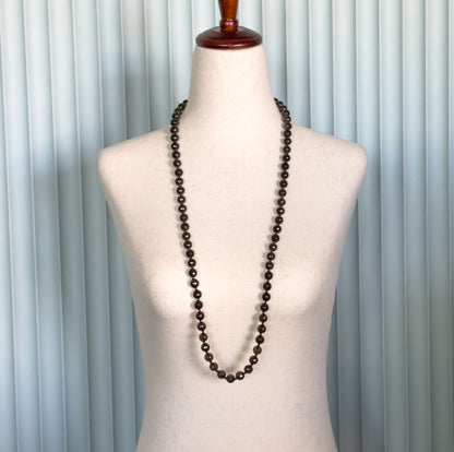 Shown at the neck on a linen dress form as one long single strand, the faceted deep brown beads falling in a slim U to below the chest.