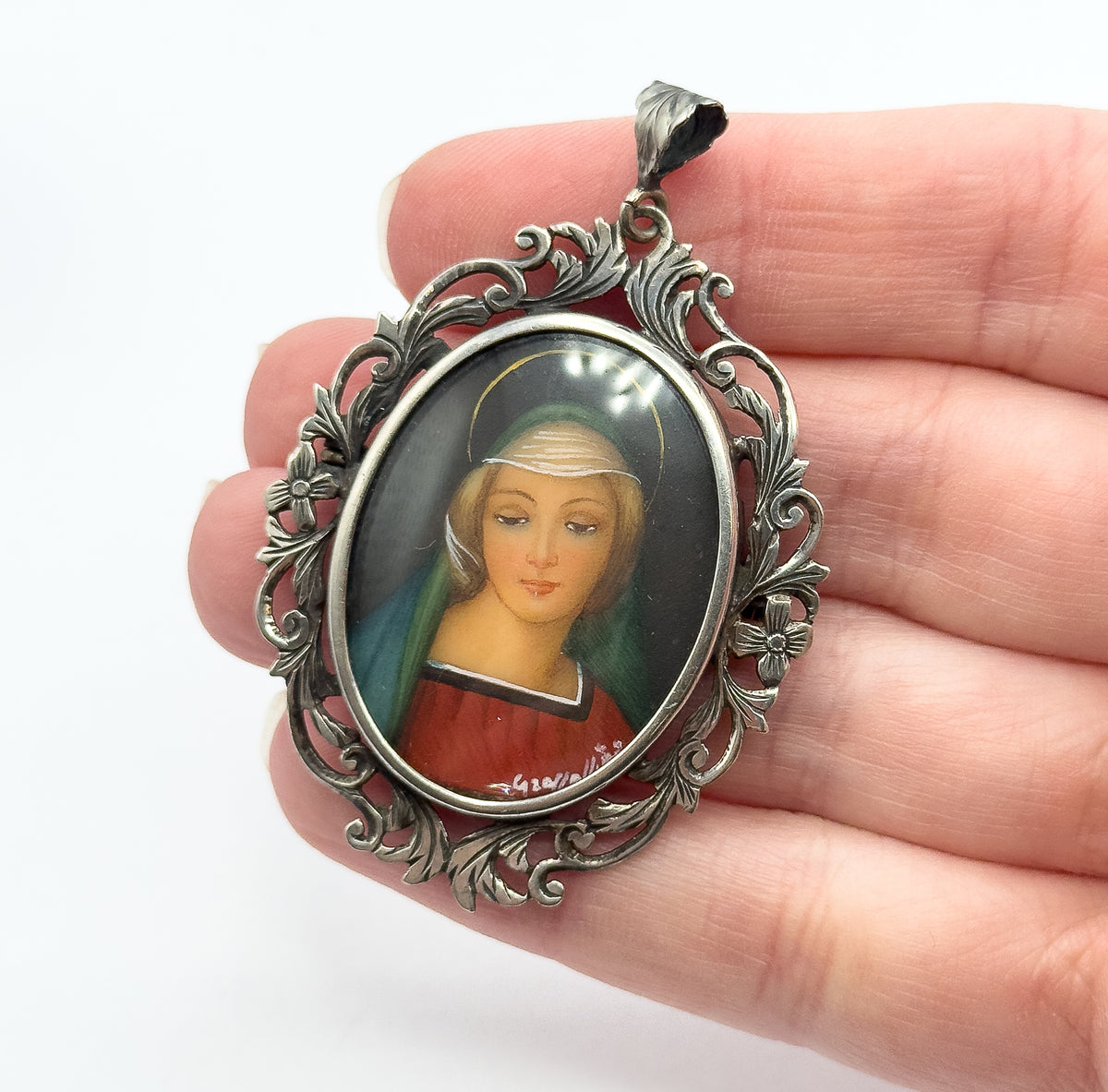 Held on open fingers, the pendant fills the width of two fingers, showing the leaf-capped bail above the acanthus scrollwork and the soft colours of the painted portrait behind glass.