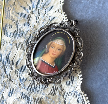 Edwardian hand-painted portrait pendant in 800 silver, shown face-up on cream lace, its oval glass-covered painting of Mother Mary framed by patinated acanthus leaf and flower scrollwork.
