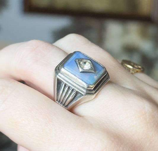 A sterling silver signet-style ring with a rectangular bluish opaline glass cabochon and round colourless paste accent, shown on a finger with stepped, ribbed shoulders in view.