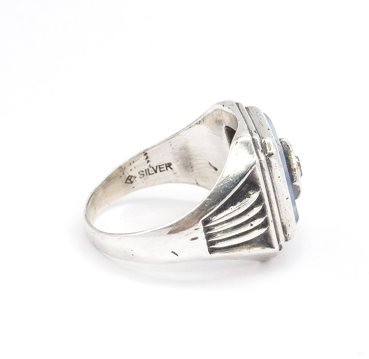 Side view of the ring resting on a white surface, showing the ribbed shoulder rising to the bezel and the stamped mark inside the shank reading SILVER beside a small maker's mark.