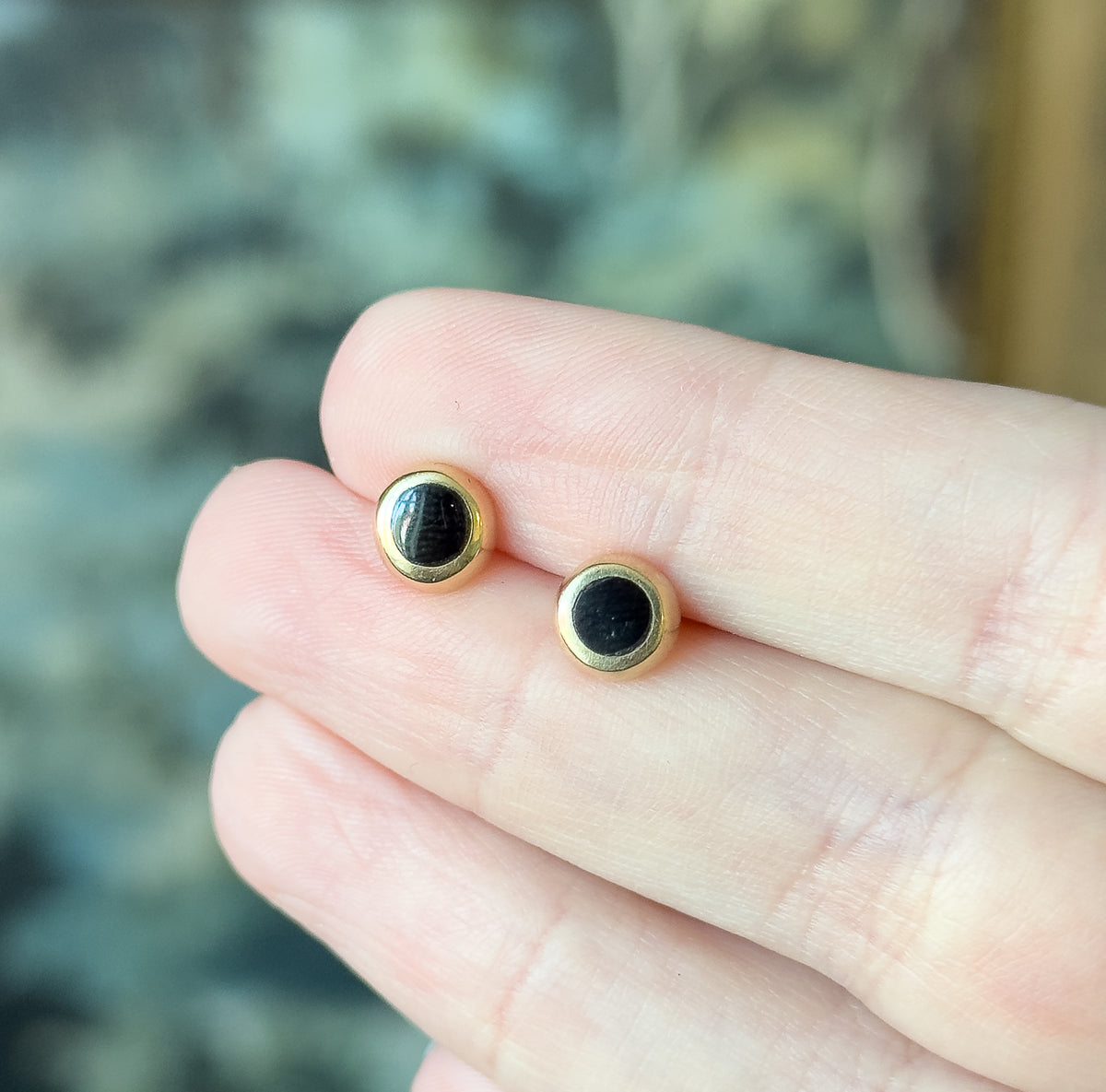 Both round black enamel studs rest on a fingertip, their polished yellow gold bezels facing the camera, showing how small the pair is against the hand.