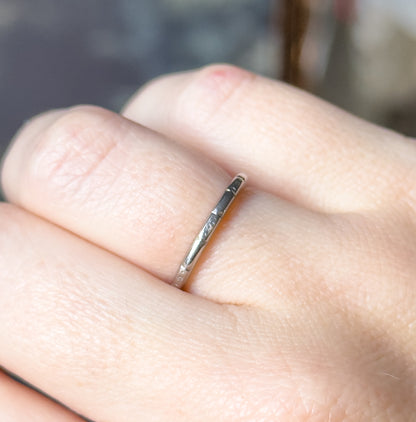 Art Deco geometric wedding band in platinum, shown on a finger in close-up, the slim etched shank seen edge-on between two fingers against a softly blurred background.
