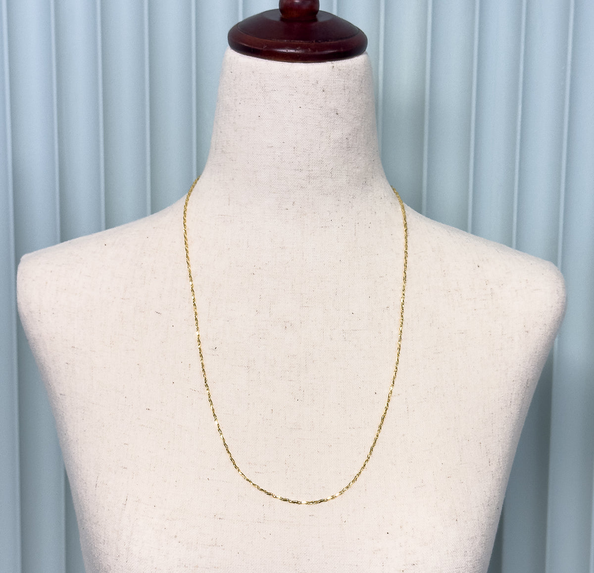 A 14K yellow gold crinkle link chain necklace shown draped at the neck of a pale linen display bust against a soft blue background.