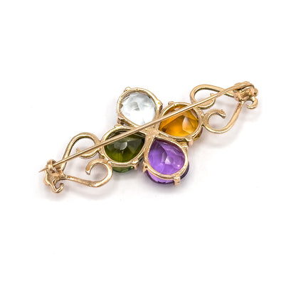 Reverse of the brooch seen from above, the open-backed settings showing the pale blue, green, yellow and purple stones through their prongs, with the pin stem crossing diagonally to the catch.