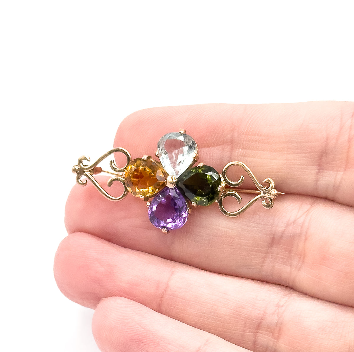 Clover brooch in 14K rose gold set with four pear-shaped stones — aquamarine, tourmaline, citrine and amethyst — between scrolled ends, shown resting on fingers to give its small scale.