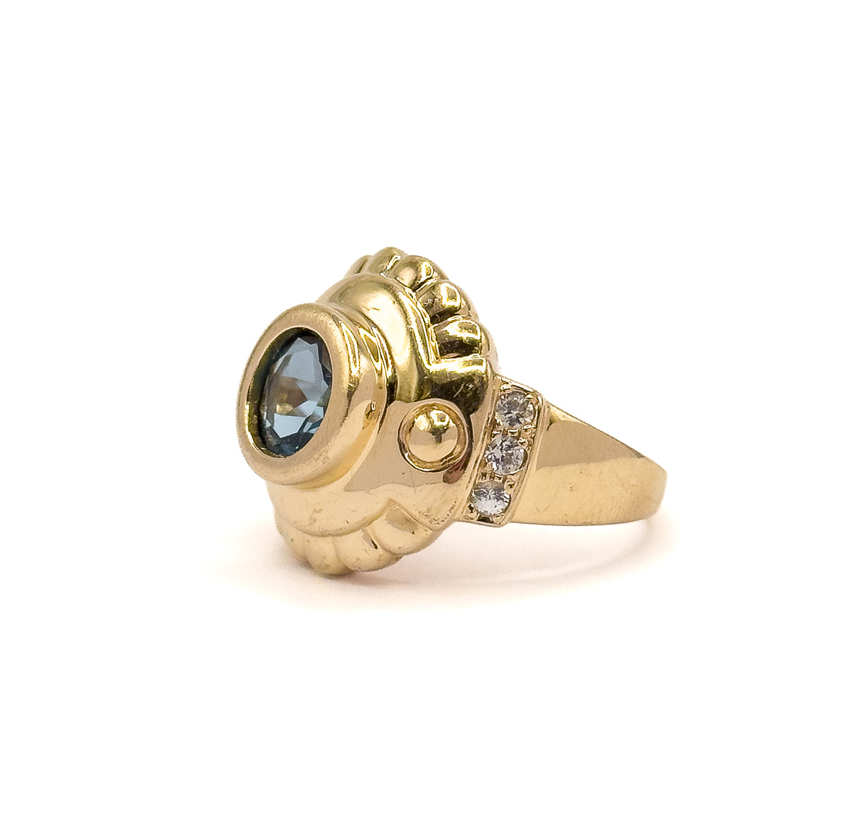 Angled three-quarter view from the front, the bezel-set London blue topaz tilted toward the viewer with the ribbed crest rising behind it and the gold bead and sapphire-set shoulder to the right.