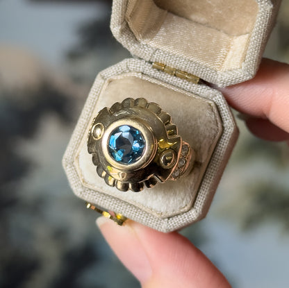 London blue topaz and white sapphire ring in 10K yellow gold, shown nestled in a small cream ring box held between finger and thumb, the bezel-set blue stone facing the camera.