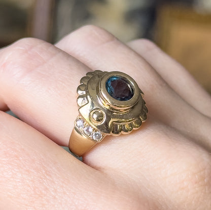 Shown on a finger at close range, the scalloped yellow gold surround and prong-set white sapphires along one shoulder visible beside the bezel-set blue topaz.
