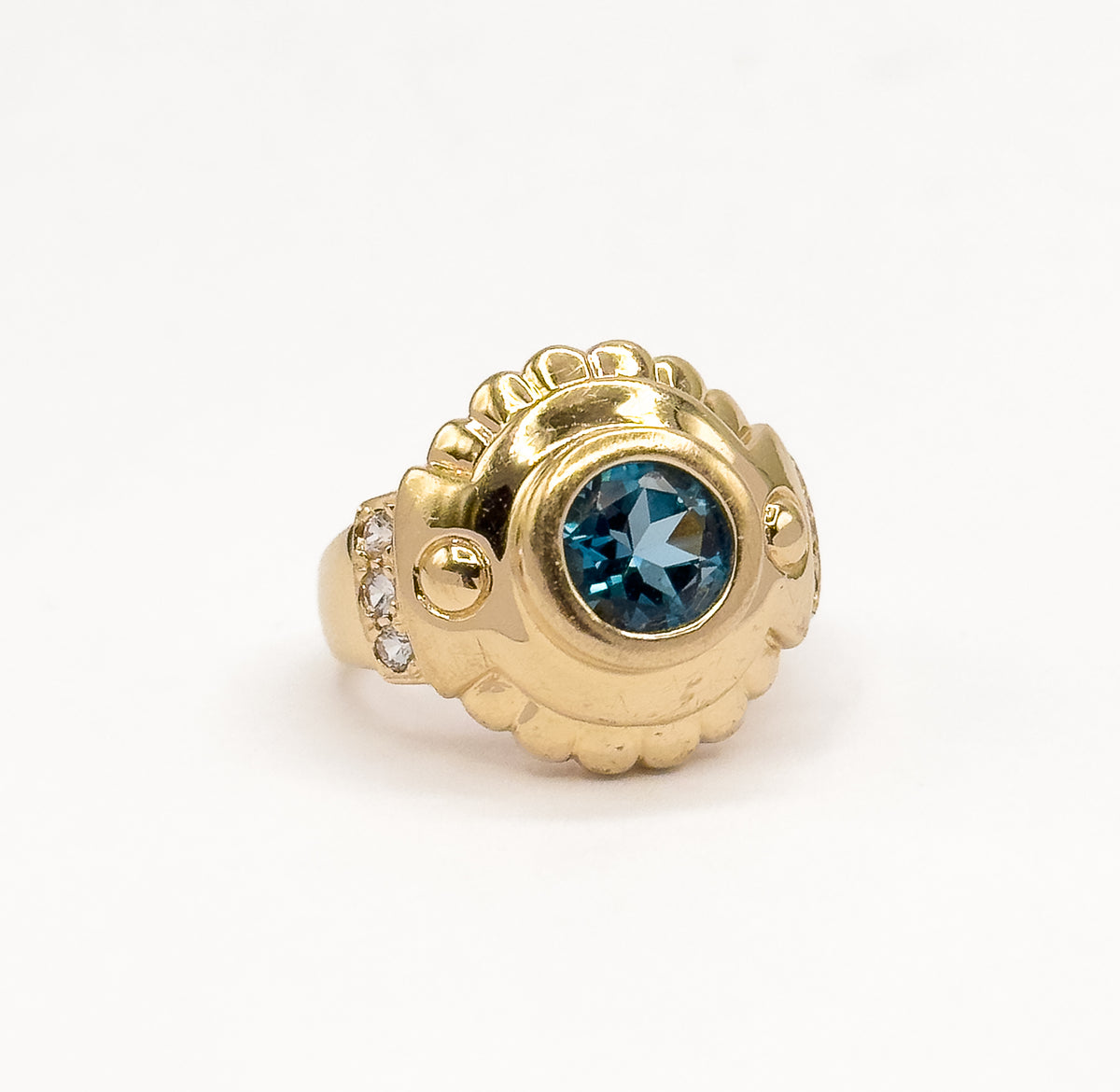 Standing upright against a plain white background, the ring seen at a three-quarter angle so the domed scalloped gold face, bezel-set blue topaz and three white sapphires on the shoulder show.