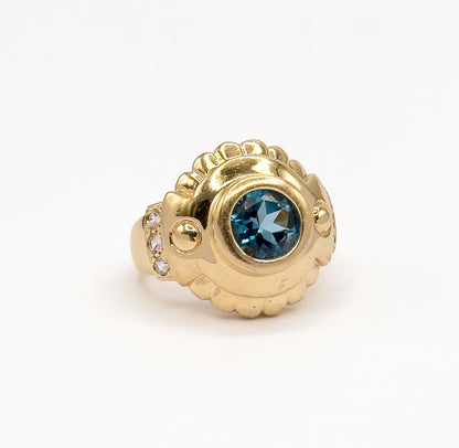 Standing upright against a plain white background, the ring seen at a three-quarter angle so the domed scalloped gold face, bezel-set blue topaz and three white sapphires on the shoulder show.