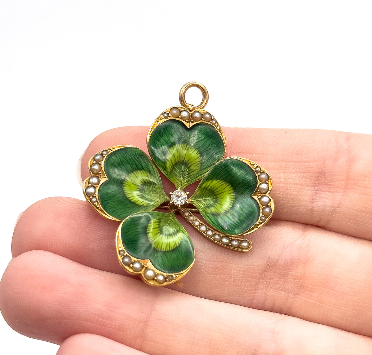 Edwardian green enamel clover brooch pendant in 14K yellow gold, seed pearls edging each leaf around a prong-set old European cut diamond, shown resting on fingertips for scale.