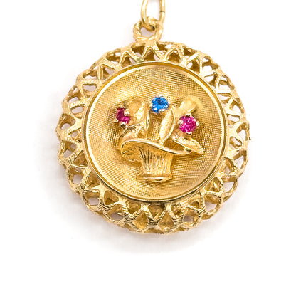 Straight-on close-up filling the frame, the round pendant centred with its bail loop at top, two red and one blue stone bright among the flowers above the textured basket, lattice rim all round.