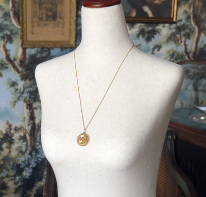 The necklace shown at the neck on a cream dress form, the fine gold chain looping down to the small oval pendant resting mid-chest against a tapestry backdrop.