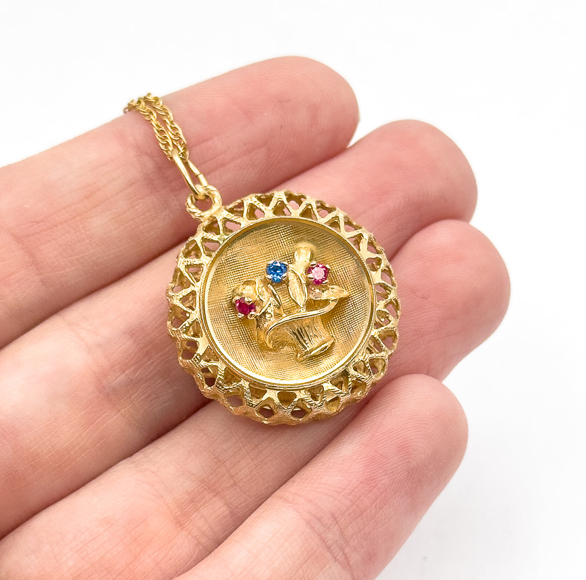 Close view of the pendant lying across the fingers with the chain trailing up over the hand, showing its size and the three small red and blue stones among the raised flowers.