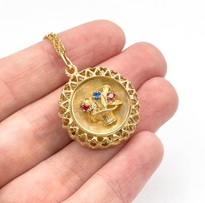 Close view of the pendant lying across the fingers with the chain trailing up over the hand, showing its size and the three small red and blue stones among the raised flowers.